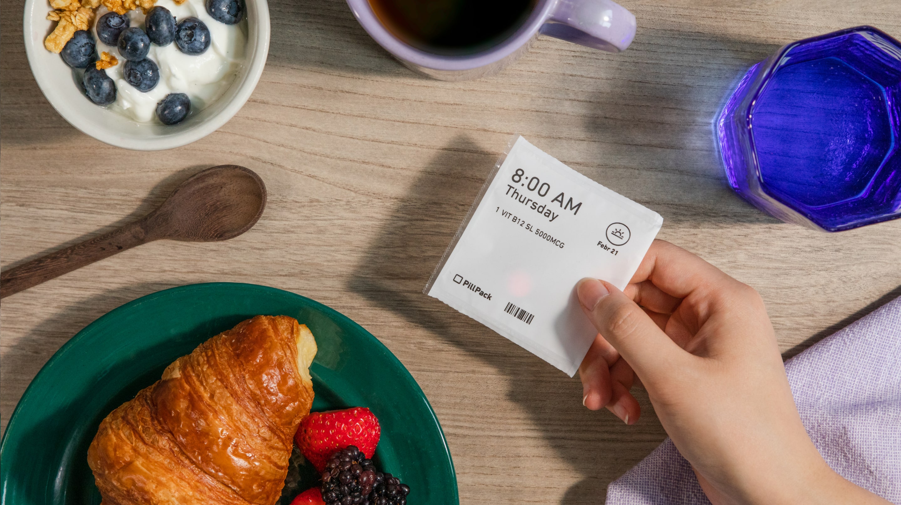 Hand holding a PillPack packet in a breakfast scene with breat, fruit, yogurt and berries, a cup of water, and coffee